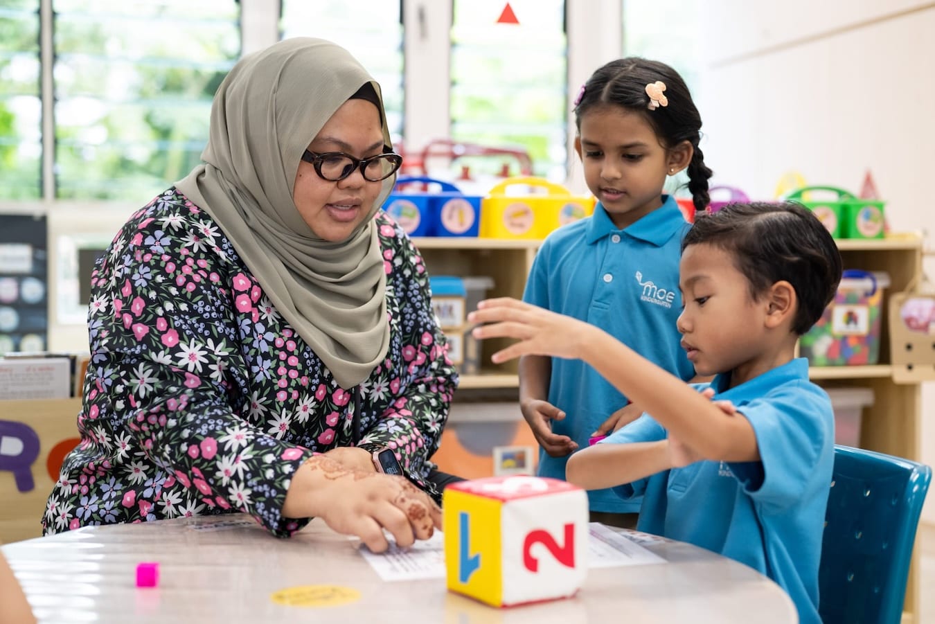 a bespectacled teacher guiding children at a table during classroom activities