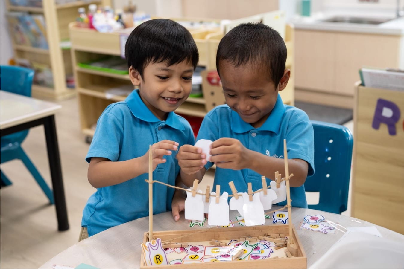two kindergarteners playing with small pieces of paper cut into shapes of clothing with alphabets on each one, and clipping them onto a miniature laundry line to form words