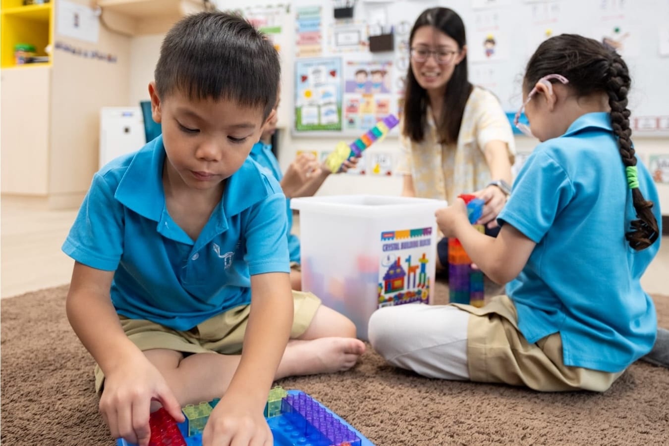 a child in the foreground plays with interlocking building blocks, while a teacher helps two other kindergarteners build their tower of blocks in the background