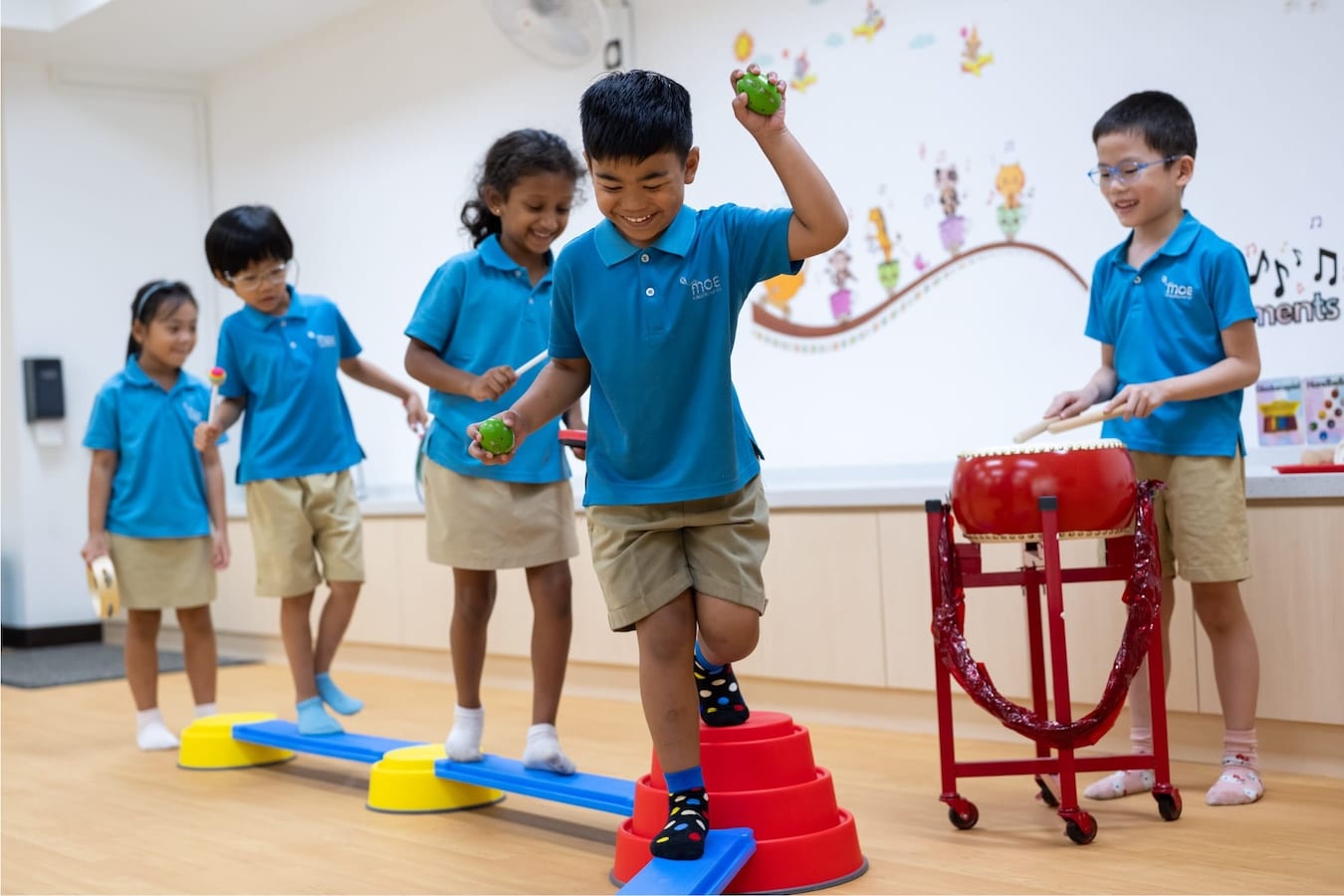 kindergarteners playing on balance beams holding little objects while one child beats on a drum