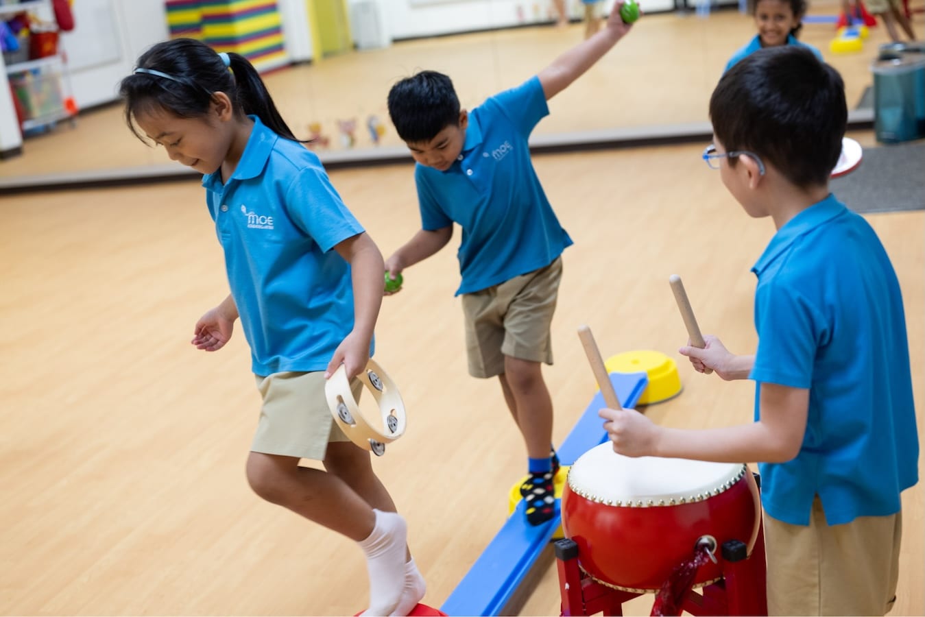 kindergarteners playing on balance beams holding little objects while one child beats on a drum